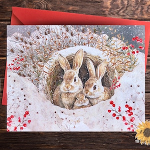 May include: Two brown rabbits peek out from a snow-covered nest with red berries and a snowy background. The rabbits have long ears and are looking at the viewer.
