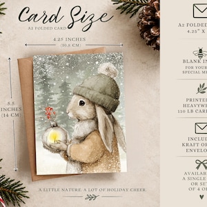 May include: An A2 folded card measuring 4.25 by 5.5 inches, featuring a watercolor illustration of a rabbit in a winter hat holding a glowing ornament. The card is printed on 110 lb cardstock, includes a kraft or red envelope, and is blank inside.