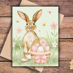 May include: Watercolor illustration of an Easter bunny with a basket of decorated eggs. The bunny is brown with long ears, standing in a field of green grass and flowers. Pink butterflies and a pink bow adorn the basket.