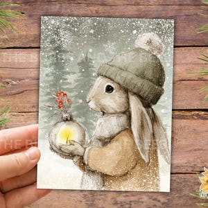 May include: A rectangular print featuring a watercolor illustration of a brown rabbit wearing a gray knit hat with a pom-pom and a scarf. The rabbit holds a glowing glass ornament topped with red berries against a snowy forest background.