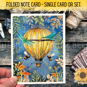 May include: Folded note card with a watercolor illustration of a hot air balloon flying through a night sky with stars, dragonflies, and bees. The balloon is yellow and white with a basket. The background is blue with green leaves and mushrooms.