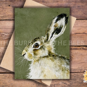 May include: A watercolor print of a hare with a green background. The hare is depicted in profile, with detailed fur and a focused gaze. The print is on a card with a brown envelope, with the text "THE BURPS AND THE BEES" and a sunflower sticker.