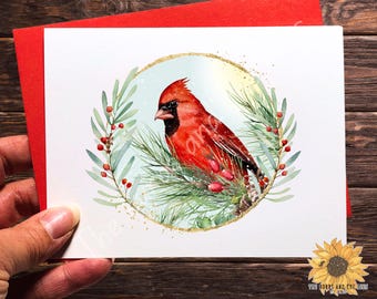 May include: A watercolor painting of a red cardinal perched on a pine branch with red berries. The bird is surrounded by a gold circle with a light blue background. The painting is on a white card with a red border.