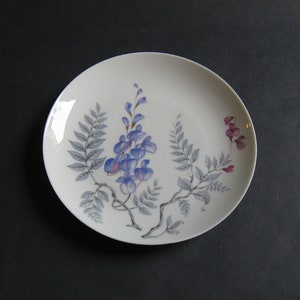 May include: A white ceramic plate featuring a floral design. The design includes blue wisteria flowers, gray leaves, and a brown stem. The plate has a gold rim and is set against a dark background.