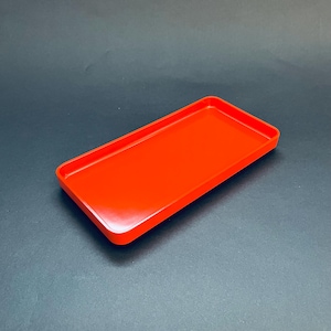 May include: A rectangular, bright red tray with slightly raised edges. The tray has a smooth, glossy finish and is empty. The tray is a solid, vibrant color and is set against a dark background.