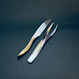May include: A stainless steel carving knife and fork set. The knife has a silver blade and a gold-toned handle. The fork has two tines and a silver finish. The set is displayed on a black background.