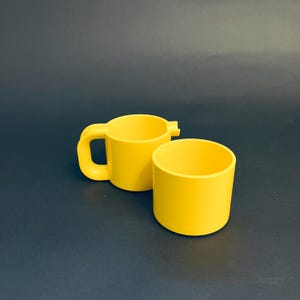 May include: Two bright yellow plastic cups. One cup has a handle and a spout-like extension. The other cup is a simple cylinder shape. Both cups are empty and sit on a dark surface.