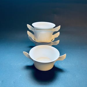 May include: Four white ceramic bowls stacked on top of each other. Each bowl has a wing-shaped handle on either side. The bowls are empty and have a glossy finish. The background is a solid dark blue.