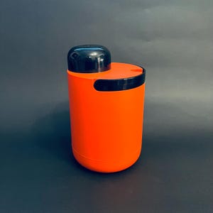 May include: An orange cylindrical container with a black lid and accents. The container has a rounded base and a curved handle-like cutout. The lid is a rounded, glossy black. The background is a solid dark gray.