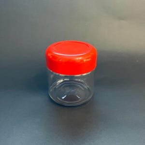 May include: A clear glass jar with a bright red lid. The jar is cylindrical and appears empty. The lid is a solid, rounded shape. The background is a dark gray color.