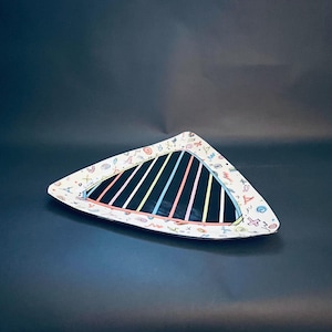 May include: A triangular decorative plate with a black center and colorful vertical stripes. The white border is adorned with small, colorful doodles. The plate is set against a dark background.
