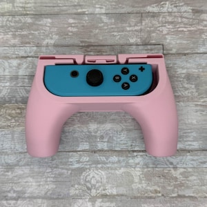 Pode incluir: Uma pega de controlador rosa com um controlador turquesa inserido. A pega tem um design curvo com uma ranhura para o controlador. O controlador tem botões pretos e um joystick. O fundo é um grão de madeira cinza claro.