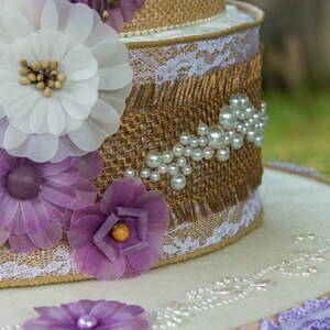 May include: A two-tiered cake decorated with burlap, lace, and white and purple fabric flowers. The cake is adorned with pearl-like beads.