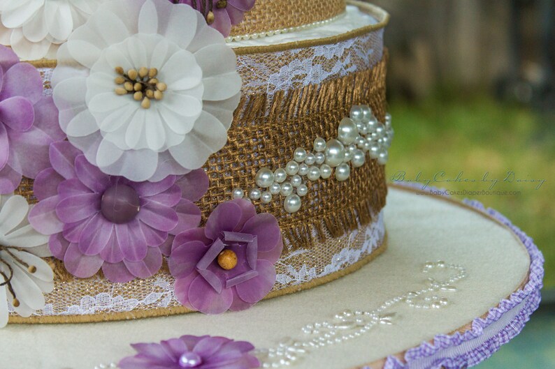 May include: A two-tiered cake decorated with burlap, lace, and white and purple fabric flowers. The cake is adorned with pearl-like beads.