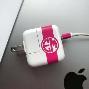 May include: White power adapter with a pink stripe and a monogram. The charger is connected to a white cable with a pink tip. The adapter is resting on a silver laptop.