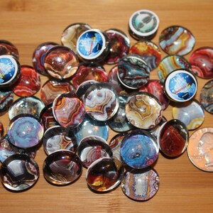 May include: A collection of colorful glass cabochons with various patterns and designs. Some of the cabochons feature images of a rocket ship, a globe, and a footprint. The cabochons are approximately 1 inch in diameter.
