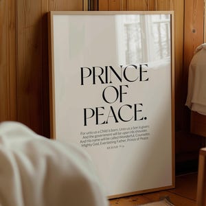 Puede incluir: Un cartel enmarcado con las palabras "PRINCE OF PEACE" en letras negras grandes y en negrita. Debajo, un texto más pequeño incluye un versículo bíblico de Isaías 9:6. El marco es de color madera clara y el fondo es blanco roto neutro.