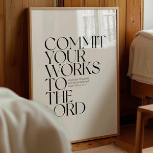 Op de afbeelding: Een ingelijste print met de woorden "COMMIT YOUR WORKS TO THE LORD" in grote zwarte letters. De tekst bevat het bijbelvers Spreuken 16:3. De print zit in een licht houten frame en leunt tegen een houten muur.