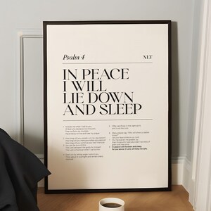 May include: A framed print with the text "IN PEACE I WILL LIE DOWN AND SLEEP" from Psalm 4. The print is in a black frame and is propped against a light blue wall. A white mug of coffee sits on a book on the floor.