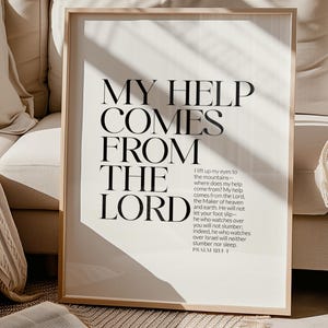 May include: A framed print with the text "MY HELP COMES FROM THE LORD" in black, set against a white background. The print includes a Bible verse from Psalm 121:1-1. The frame is a light wood color. The print is displayed on a beige sofa.