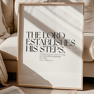 May include: A framed print with the text "THE LORD ESTABLISHES HIS STEPS." The quote from Proverbs 16:9 is in a minimalist black font on a white background. The print is in a light wood frame, resting on a beige rug near an open book.