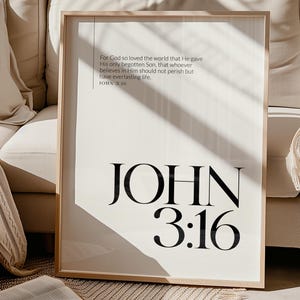May include: A framed print featuring the text "JOHN 3:16" in a large, bold font. The print also includes a Bible verse in a smaller font. The frame is light-coloured, and the background is a neutral white.