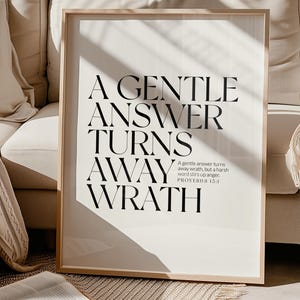 Può includere: Una stampa incorniciata con il testo "A GENTLE ANSWER TURNS AWAY WRATH" in nero su sfondo bianco. La stampa è in una cornice di legno chiaro ed è appoggiata a un divano color crema. Un libro aperto è davanti alla stampa.