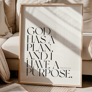 Può includere: Una stampa incorniciata con il testo "GOD HAS A PLAN, AND I HAVE A PURPOSE." in nero su sfondo bianco. La stampa è in una cornice di legno chiaro ed è esposta vicino a un divano e a un libro aperto.