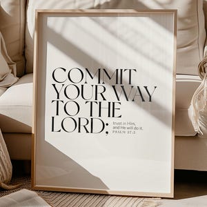 May include: A framed print with the text "COMMIT YOUR WAY TO THE LORD: trust in Him, and He will do it. PSALM 37:5" in black font. The print is in a light wood frame, resting against a beige sofa. An open book and a beige rug are in the foreground.
