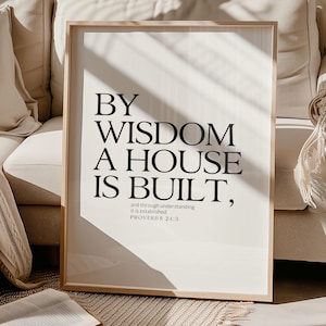 Pode incluir: Uma impressão emoldurada com o texto "BY WISDOM A HOUSE IS BUILT" em preto, com o versículo bíblico Provérbios 24:3. A impressão está em uma moldura de madeira clara, repousando sobre um tapete com um livro aberto na frente.