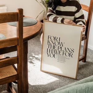 May include: A beige poster with a black and white quote that reads "FOR I KNOW THE PLANS I HAVE FOR YOU" and a smaller quote from Jeremiah 29:11. The poster is framed in a thin, gold frame.