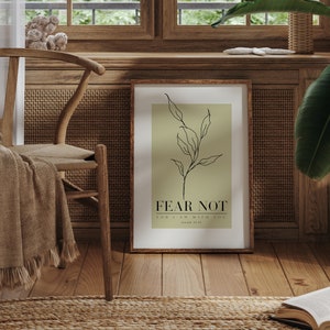 May include: A framed print with a green background and a black line drawing of a branch with leaves. The text "FEAR NOT" is printed in black below the drawing, followed by "FOR I AM WITH YOU" in smaller font. The print is hanging on a wall in a room with a wooden chair and a rug.