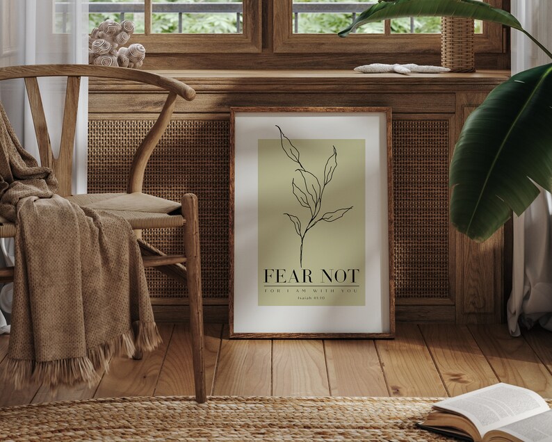 May include: A framed print with a green background and a black line drawing of a branch with leaves. The text "FEAR NOT" is printed in black below the drawing, followed by "FOR I AM WITH YOU" in smaller font. The print is hanging on a wall in a room with a wooden chair and a rug.