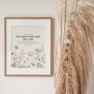 May include: A framed print with a white background and a botanical illustration of wildflowers. The text "I AM THE Resurrection and the Life" is printed in black at the top of the print. The scripture reference "John 11:25" is printed below the text.