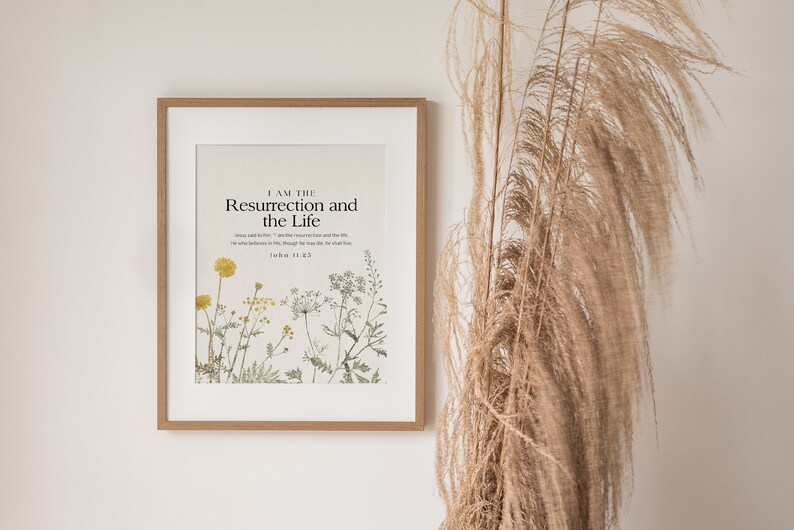 May include: A framed print with a white background and a botanical illustration of wildflowers. The text "I AM THE Resurrection and the Life" is printed in black at the top of the print. The scripture reference "John 11:25" is printed below the text.