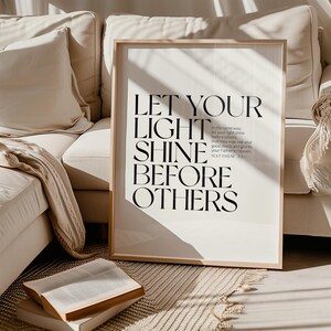 May include: A framed print with the text "LET YOUR LIGHT SHINE BEFORE OTHERS" in black against a white background. The print is in a light wood frame and is displayed near a beige sofa and a book.