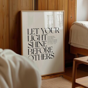 May include: A framed poster with the text "LET YOUR LIGHT SHINE BEFORE OTHERS" in black against a cream background. The poster includes a Bible verse from Matthew 5:16. The frame is a light wood color, and the poster is displayed in a bedroom setting.