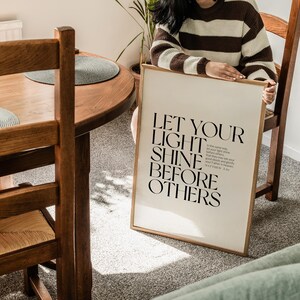 May include: A framed print with the text "LET YOUR LIGHT SHINE BEFORE OTHERS" in black. The print is in a light wood frame and is held by a person wearing a brown and cream striped sweater. The print is leaning against a wooden chair.