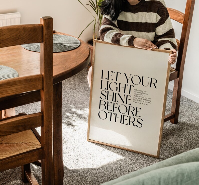 May include: A framed print with the text "LET YOUR LIGHT SHINE BEFORE OTHERS" in black. The print is in a light wood frame and is held by a person wearing a brown and cream striped sweater. The print is leaning against a wooden chair.