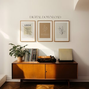May include: Three framed prints with minimalist line drawings of branches and leaves. The prints are hung on a white wall above a wooden cabinet with a record player on top. The prints have the text "ABIDE", "HIS MERCIES ARE NEW EVERY MORNING", and a simple leaf design.