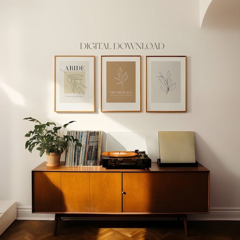 May include: Three framed prints with minimalist line drawings of branches and leaves. The prints are hung on a white wall above a wooden cabinet with a record player on top. The prints have the text "ABIDE", "HIS MERCIES ARE NEW EVERY MORNING", and a simple leaf design.