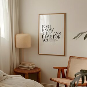 May include: A framed print with a white background and black text that reads "For I know the plans I have for you." The text is a quote from the Bible, Jeremiah 29:11. The print is hanging on a light beige wall in a room with a wooden chair and a lamp.
