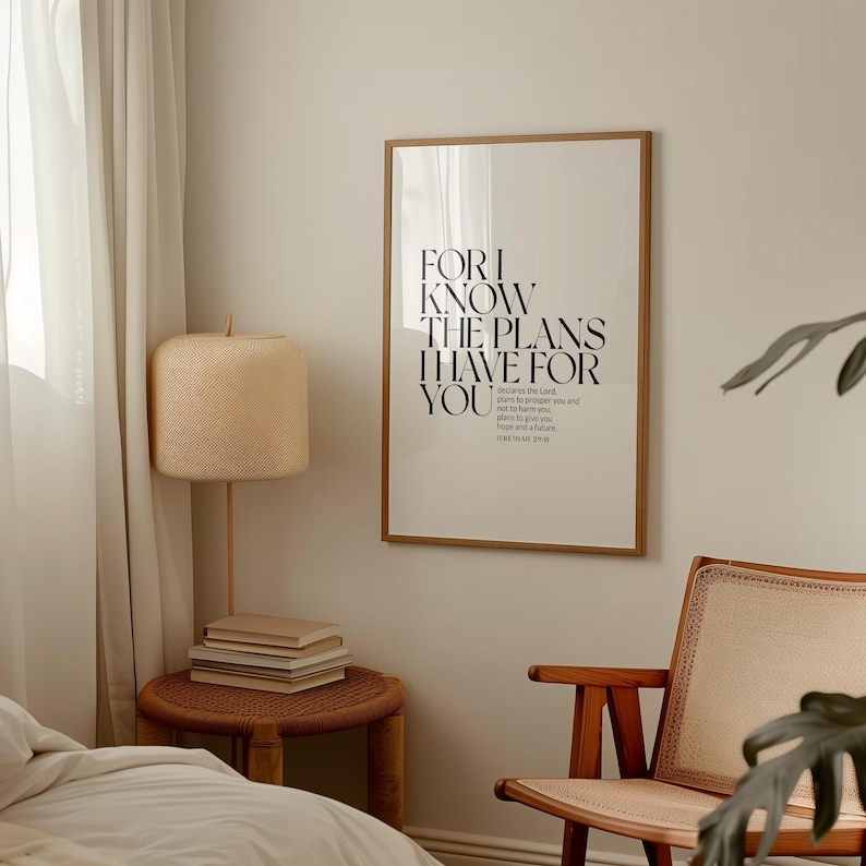 May include: A framed print with a white background and black text that reads "For I know the plans I have for you." The text is a quote from the Bible, Jeremiah 29:11. The print is hanging on a light beige wall in a room with a wooden chair and a lamp.