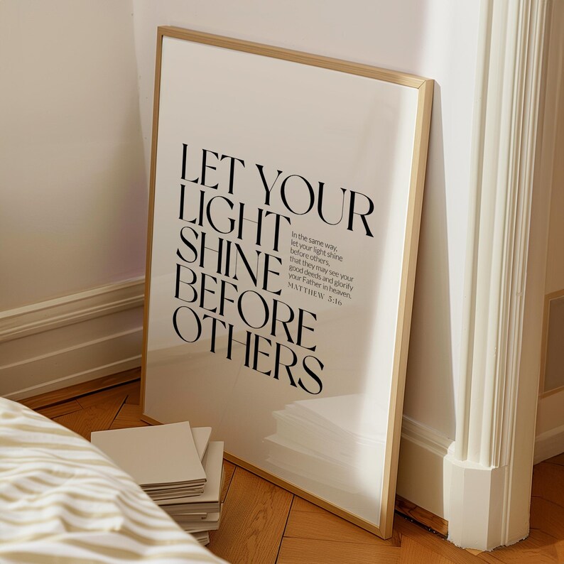 May include: A framed print with the words "LET YOUR LIGHT SHINE BEFORE OTHERS" in black font. The print has a gold-colored frame and is leaning against a white wall. The text includes a Bible verse from Matthew 3:16.