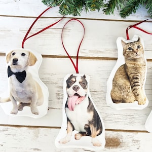 Peut inclure: Ornements de No&euml;l avec des portraits d'animaux de compagnie. Les d&eacute;corations comprennent un chien avec un n&oelig;ud papillon, un bouledogue avec la langue sortie et un chat tigr&eacute;. Chaque ornement a un ruban rouge pour l'accrochage, sur un fond en bois blanc.