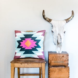 May include: A wooden chair with a colorful geometric patterned pillow on the seat. The chair is next to a stack of wooden crates, one of which has the text "Svea Margarin" printed on it. A white cow skull with horns is mounted on the wall above the crates.