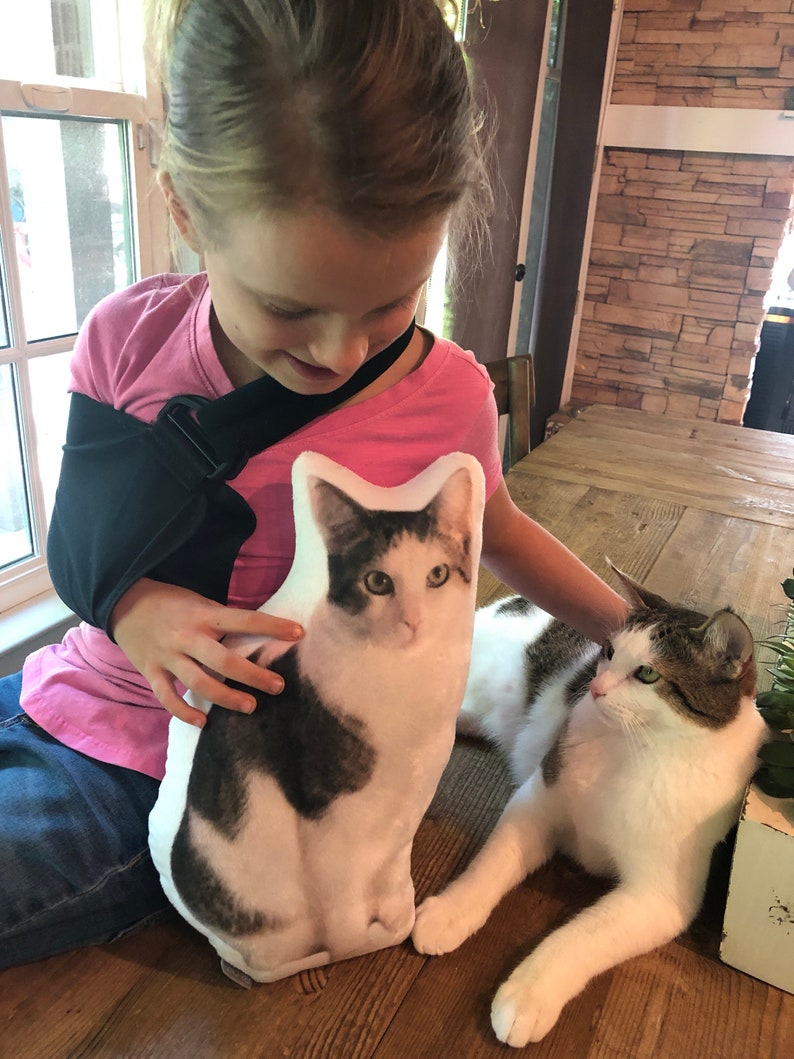 May include: A young girl wearing a pink shirt and a black sling is holding a white and grey cat cushion. A real cat with white and grey fur is lying on the floor next to the girl.