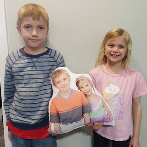 May include: Two children are holding a white cushion with a photo of two children on it. The cushion has a cloud with a smiley face on it. The children in the photo are wearing colourful shirts. The children holding the cushion are wearing striped and pink shirts.