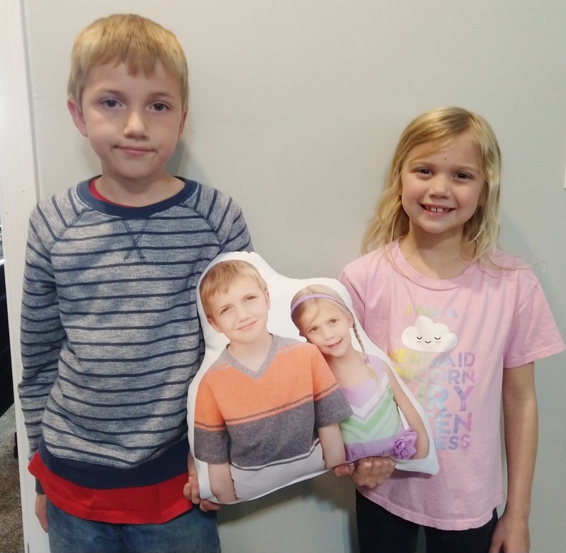 May include: Two children are holding a white cushion with a photo of two children on it. The cushion has a cloud with a smiley face on it. The children in the photo are wearing colourful shirts. The children holding the cushion are wearing striped and pink shirts.
