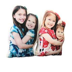 May include: Two custom photo pillows, each featuring a pair of smiling girls. One pillow shows girls in blue dresses, the other in red floral dresses. The pillows are cut in the shape of the girls hugging.